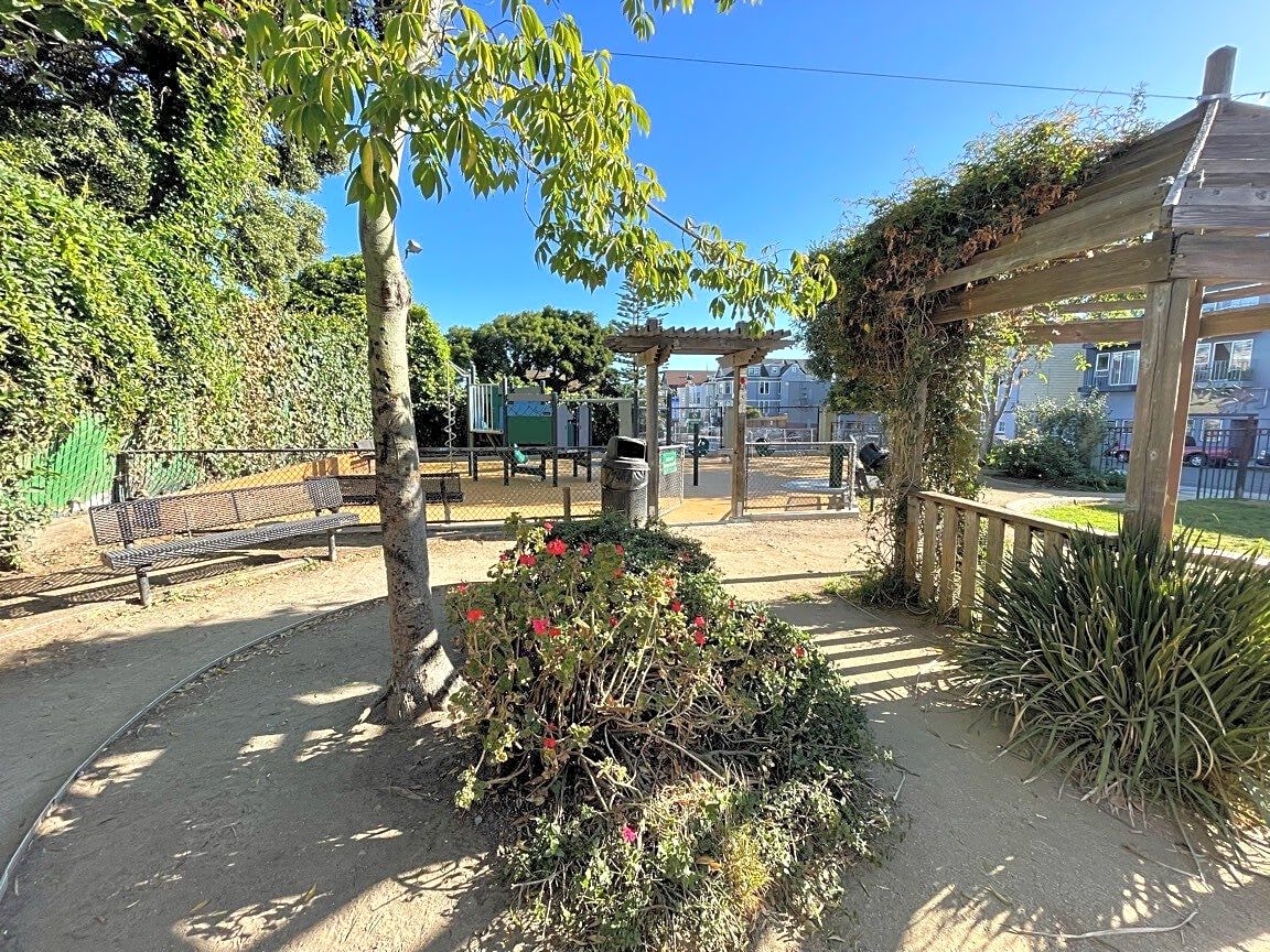 A corner of the Alioto Mini Park playground with a teal slide on rubberized safety surfacing, next to a tall, well-trimmed hedge along the fence line.